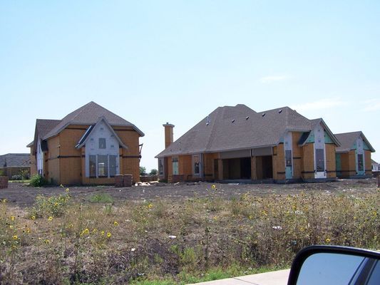 Three houses under construction, featuring tan exterior walls and brown roofs, sit on a clear, sunny day.