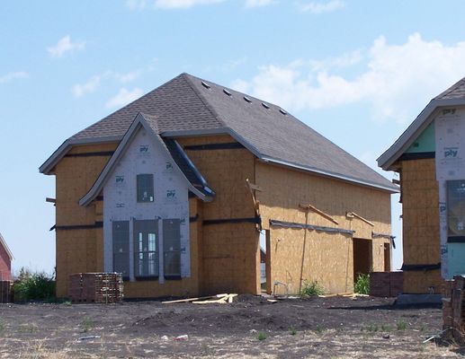 A partially constructed house frame with tan plywood siding and a gray roof under a clear blue sky.