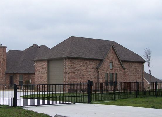 A single-story brick house with a dark roof and a black iron gate across the driveway under an overcast sky.