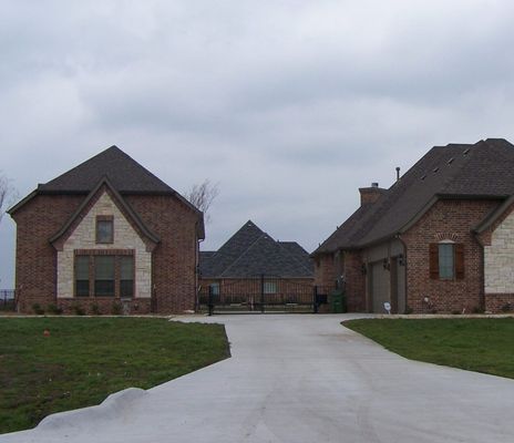 A paved driveway leads between two brick houses toward a gated entrance with another house visible in the distance.