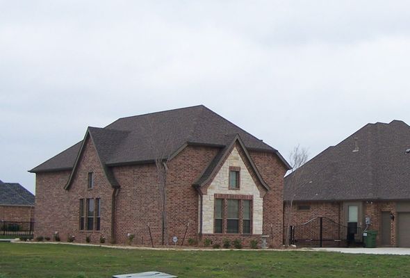 A suburban brick house with a dark shingled roof and stone accents, set against a cloudy sky on a grassy lot.