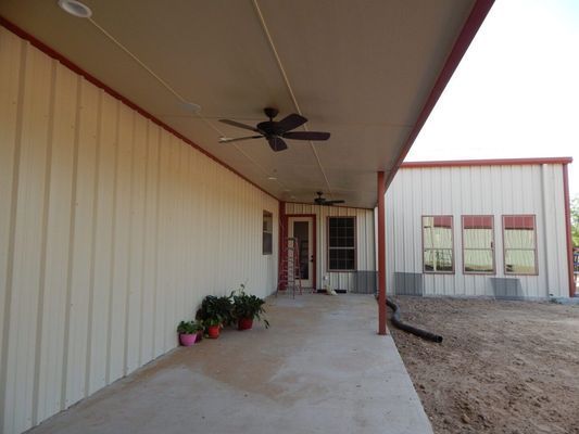 A beige metal building features a long covered porch with ceiling fans, a glass-paned door, and potted plants.