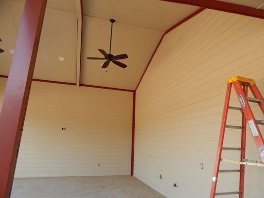An interior view of a large, high-ceilinged room with tan walls, red trim, a ceiling fan, and an orange ladder.