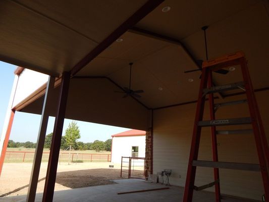 A red ladder stands in an open-air garage with brown ceilings, ceiling fans, and a view of a rural property outside.