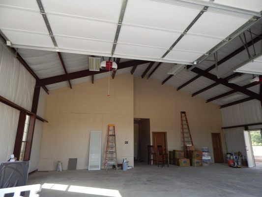 Interior view of a large, high-ceilinged workshop or garage with beige walls, exposed beams, ladders, and open doorways.
