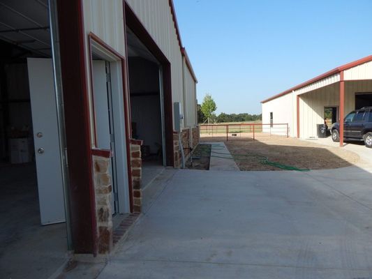 An outdoor view of two metal buildings with concrete driveways, stone accents on the walls, and a parked SUV.