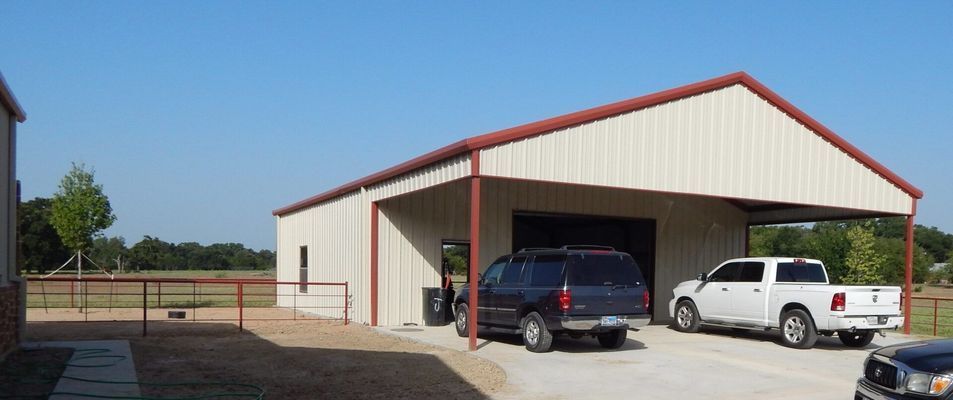 A metal garage with a covered parking area holding a dark SUV and a white truck in a rural setting with a fence.