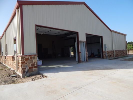 A tan metal building with stone siding and two open garage bays on a concrete driveway under a clear blue sky.