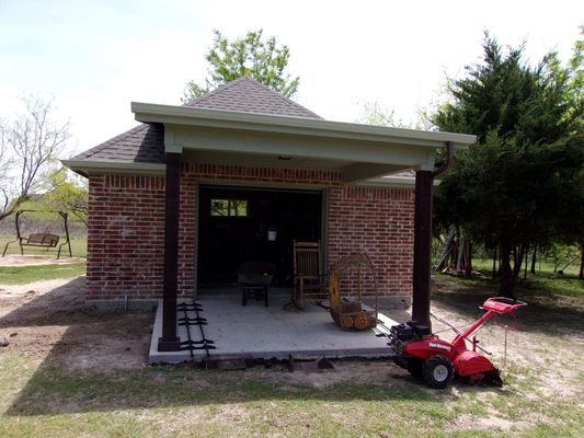 A small brick structure with a porch and hipped roof sits on a grassy lot with a red tiller parked in front.