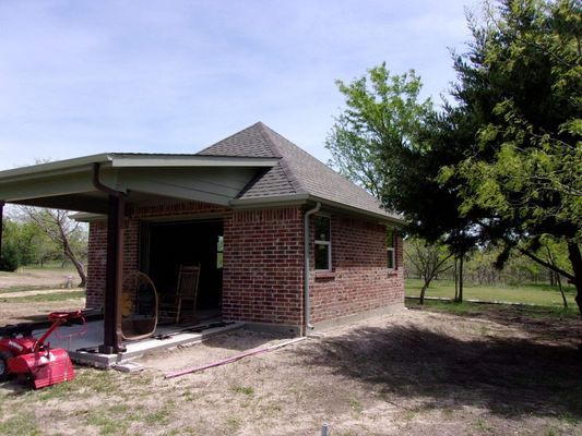 A brick outbuilding with a gray roof and an attached side porch, situated in a grassy yard on a sunny day.