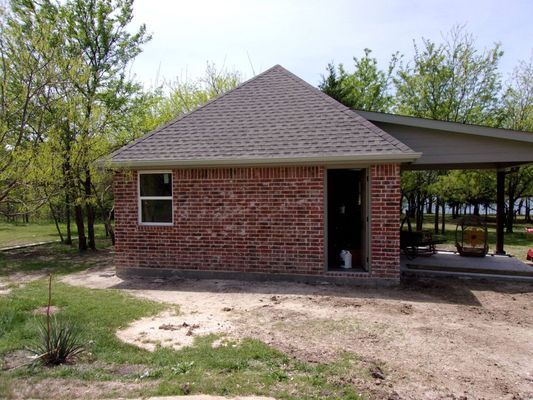 A red brick building with a hip roof and an attached side porch, situated on a grassy lot surrounded by green trees.