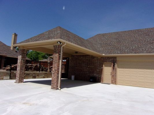 A tan brick house exterior featuring a large covered parking area with brick pillars and a light-colored concrete driveway.