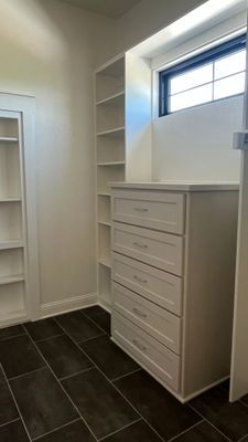 White built-in dresser and open shelving under a window in a closet with dark tiled floors.