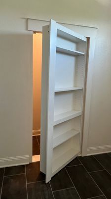 A white, shelf-lined hidden door stands slightly ajar, revealing a small, tiled interior space beyond.