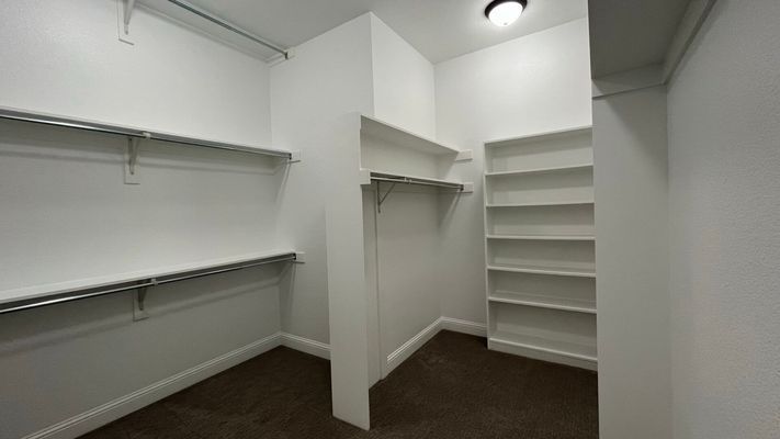 A walk-in closet with white walls, dark carpet, metal shelving rods, and a tall built-in white shelving unit.