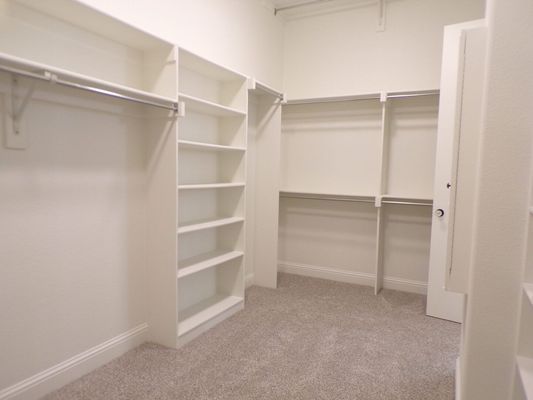 A walk-in closet featuring white shelves, hanging rods, and beige carpet.