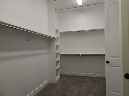 A white walk-in closet featuring built-in shelves, hanging rods, and grey carpeting, viewed from an open doorway.
