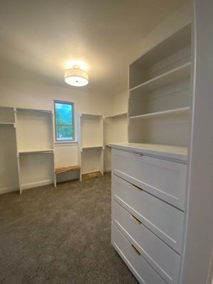 A walk-in closet featuring white wooden shelving, a built-in dresser with gold pulls, grey carpeting, and a small window.