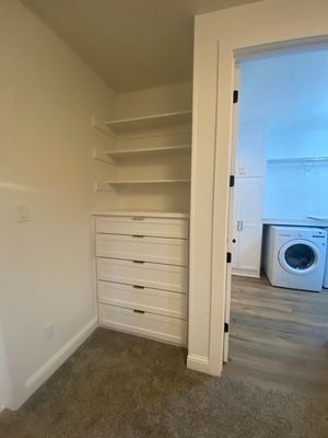 A white built-in unit with open shelves above a dresser of five drawers, located next to a doorway showing a laundry room.