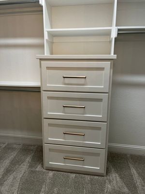 A white closet storage unit featuring four stacked drawers with brass-colored handles, situated in a carpeted walk-in closet.