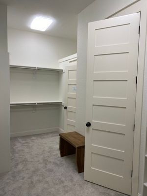 A walk-in closet featuring light gray carpet, white walls, built-in shelving, a wooden bench, and white shaker-style doors.