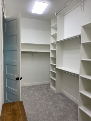 A walk-in closet featuring white shelves, hanging rods, gray carpet, a light blue door, and a wooden bench.