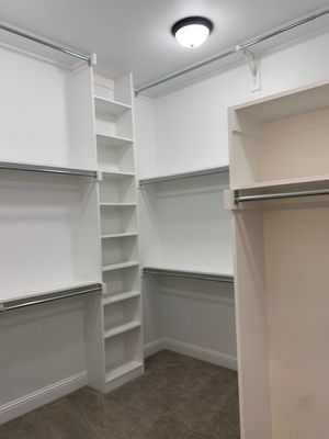 A walk-in closet featuring white wooden shelving and hanging rods against white walls over a brown carpeted floor.