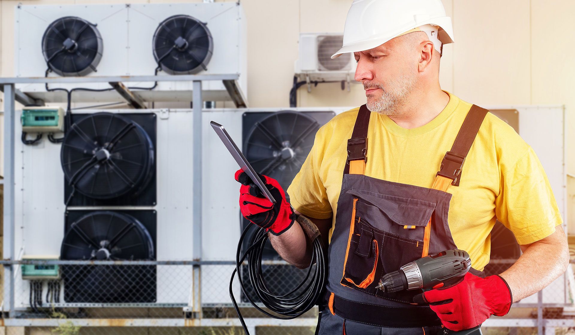 A man holding a drill and a tablet with commercial HVAC system behind hin