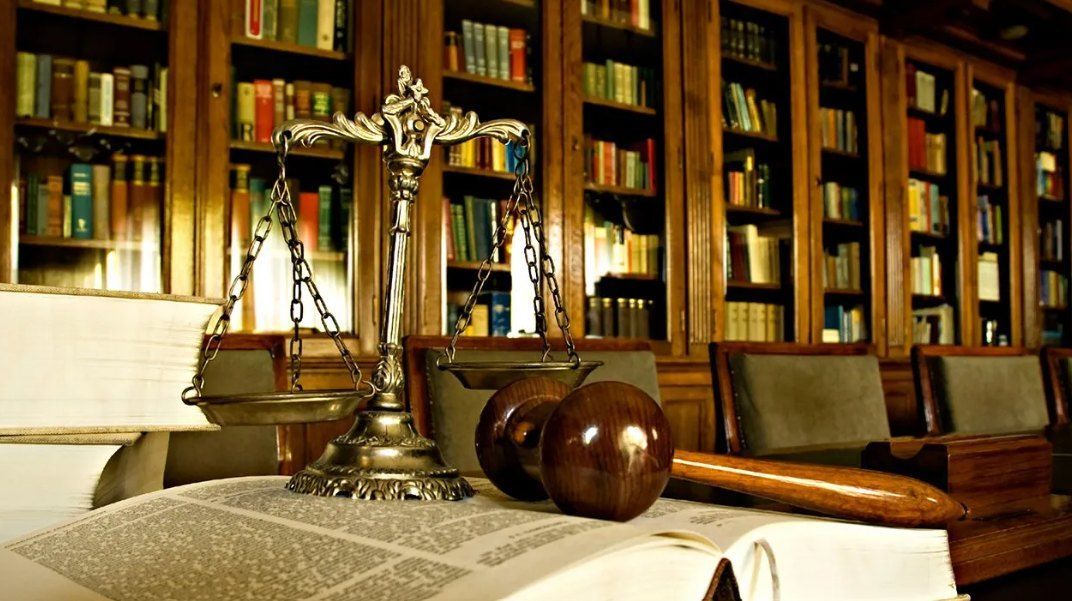 A silver scale of justice and a wooden gavel sit on a book-covered table in a wood-paneled law library.