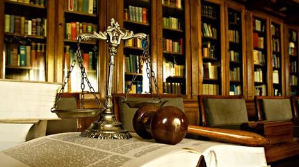 A judge’s gavel and a metallic scale of justice rest on an open book in a dimly lit law library with shelves of books.