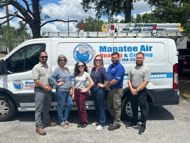 Five people standing in front of a Manee Air HVAC service van outdoors under trees.