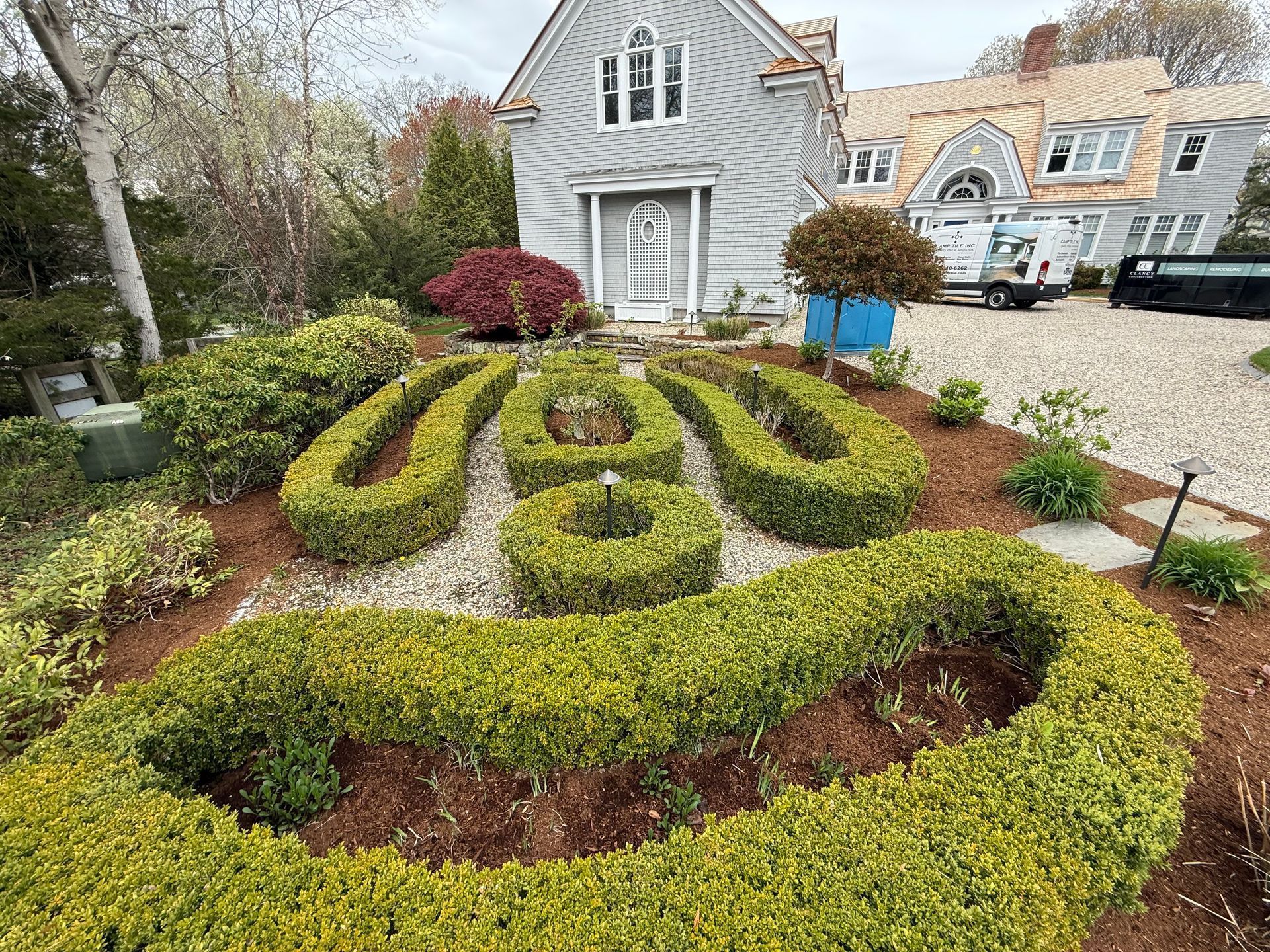 Curved boxwood garden maze in front of a gray house with stone driveway and spring flowers