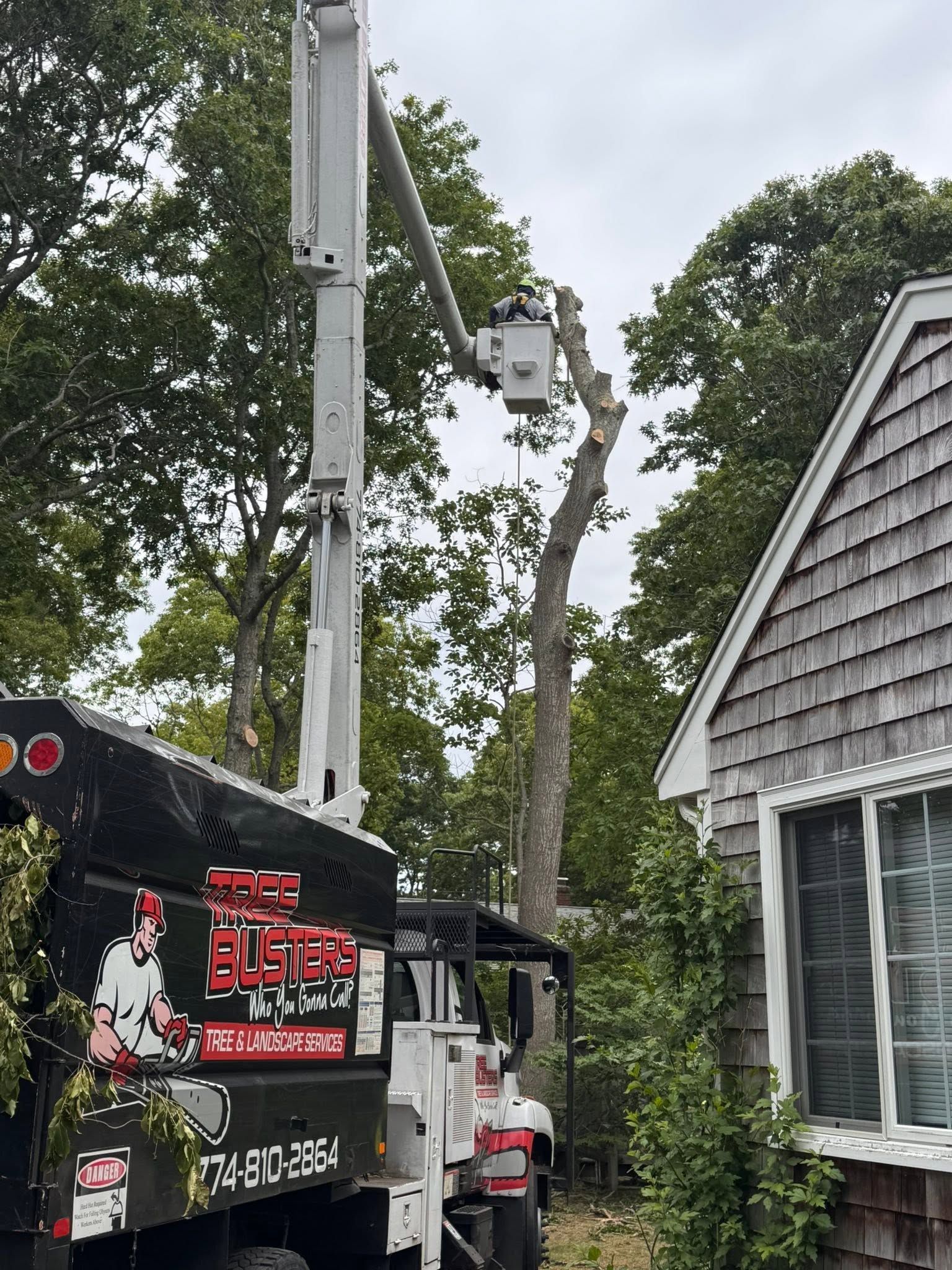 Utility truck with boom lift beside a house and tall trees, with a worker near the truck.