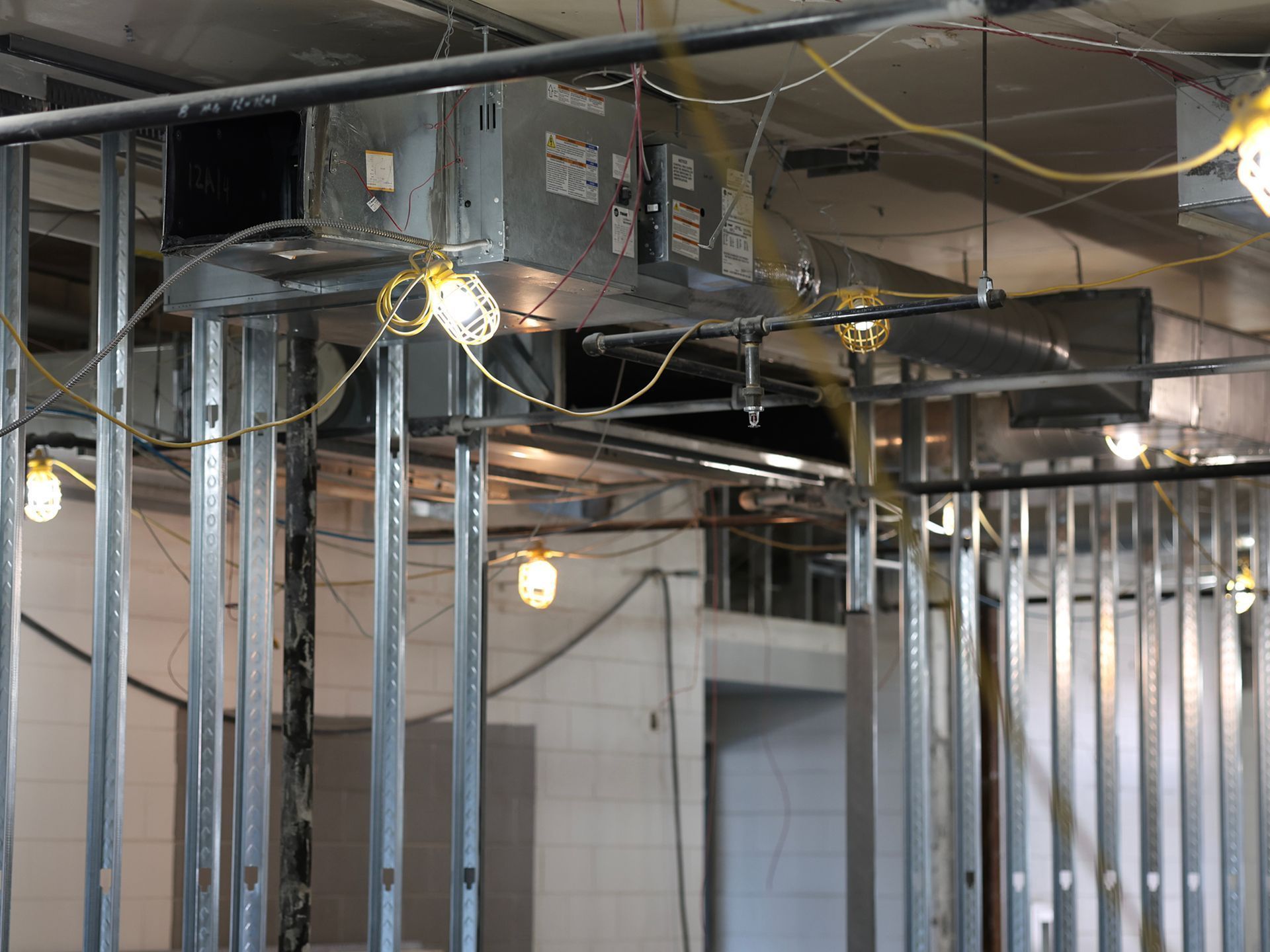 Interior construction site showing metal wall studs, ductwork, and hanging temporary work lights.
