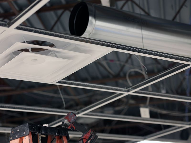 A worker on a ladder installs or repairs a white HVAC ceiling vent near exposed metal ductwork in an open ceiling.