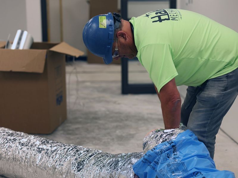 A worker in a blue hard hat and neon yellow shirt connects flexible foil ductwork inside a building under construction.