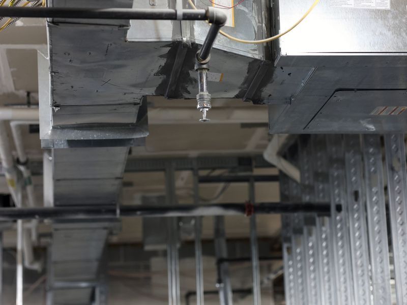 A sprinkler head pointing downward from a black pipe near metallic HVAC ductwork in a room under construction.