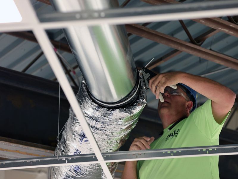A person in a bright green shirt wears safety glasses while installing metal ductwork in a ceiling grid.