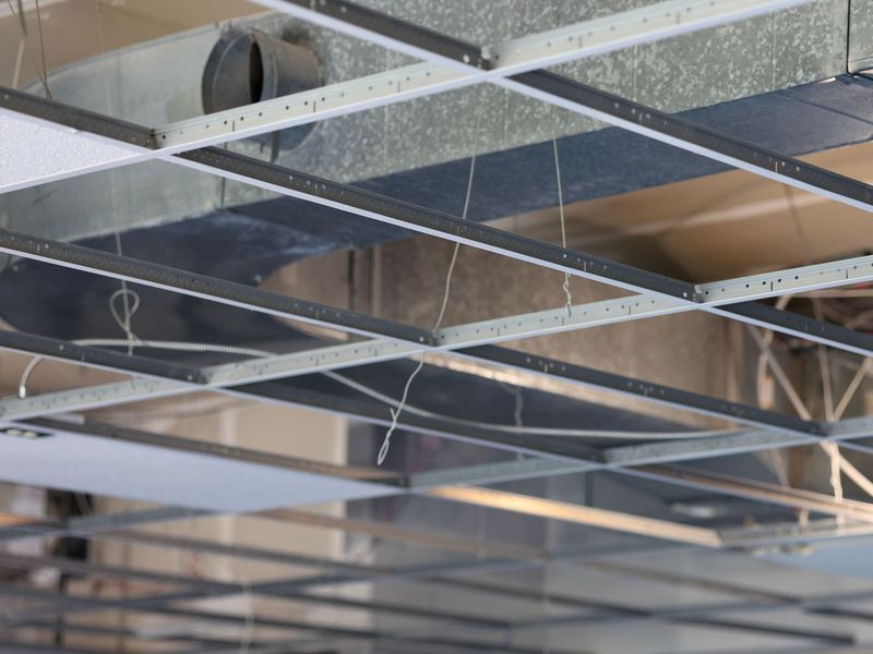 A low-angle view of a suspended drop-ceiling grid being installed, showing metal framing, ductwork, and dangling wires.