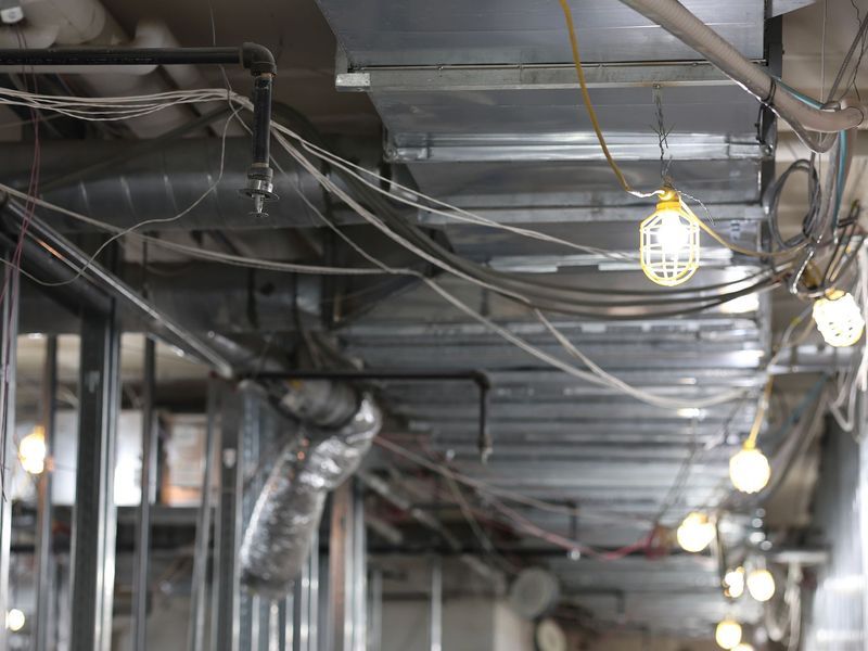 Construction site interior featuring exposed ductwork, pipes, dangling work lights, and unfinished metal framing.