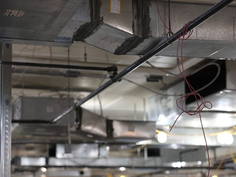Exposed ceiling infrastructure featuring metal ventilation ducts, piping, and loose red wire bundles.