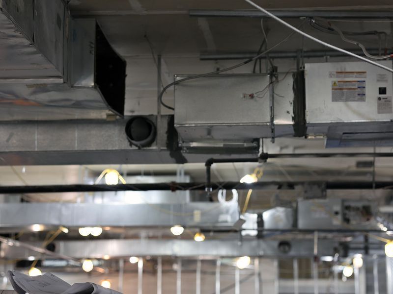 Exposed metal ventilation ductwork and pipes hang from the ceiling in an industrial building space under construction.