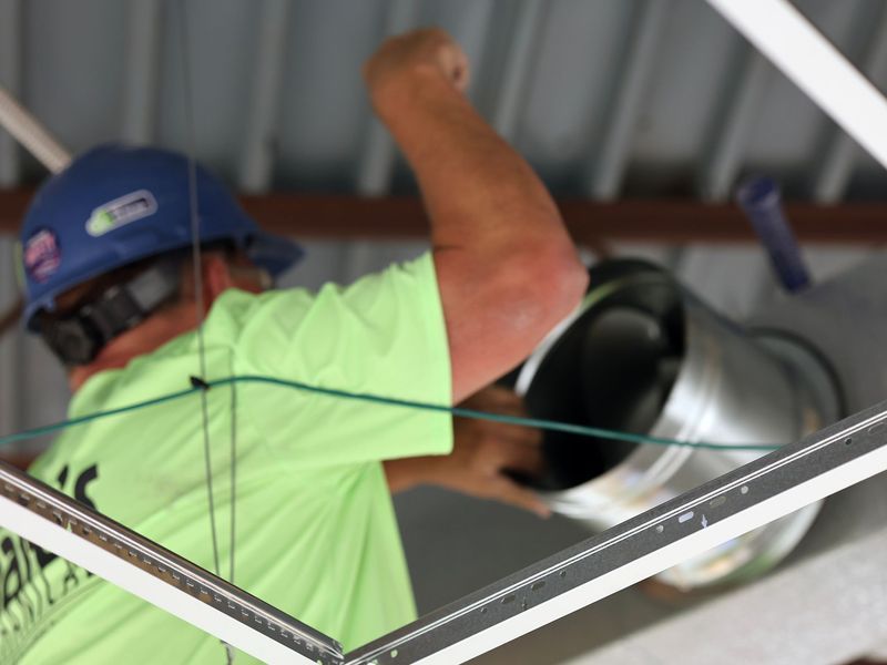 A construction worker in a lime-green shirt and blue hard hat installs circular metal ductwork in a ceiling.
