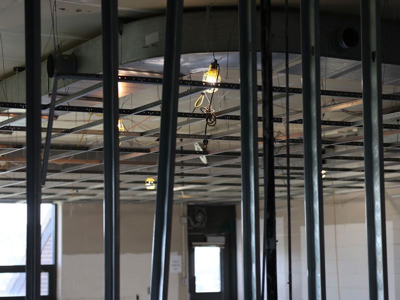 Interior view of a building under renovation, showing exposed metal ceiling framing, hanging work lights, and wall studs.