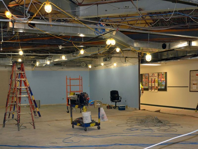Interior of an unfinished room with a ladder, rolling cart, black office chair, and exposed ceiling with hanging lights.