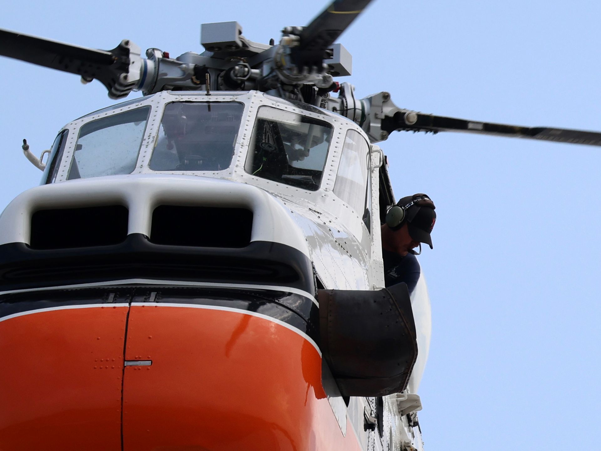 A crew member looks out from the open side door of a white and orange search and rescue helicopter against a blue sky.