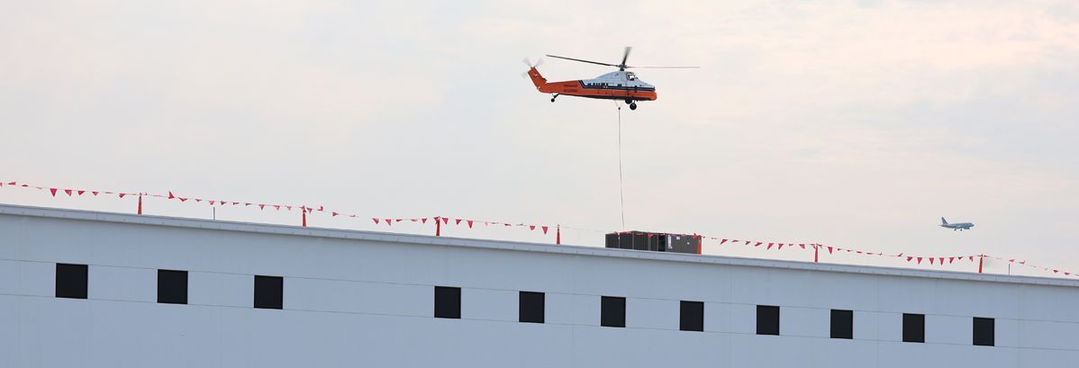 An orange and white helicopter lifts a large box onto the roof of a long, white building lined with many windows.