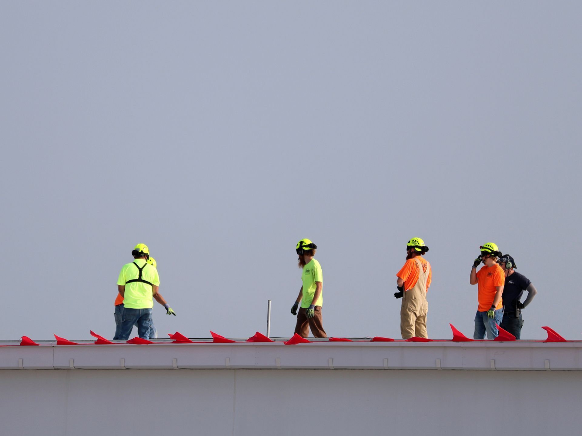 Five workers in high-visibility vests and safety helmets stand on a rooftop lined with small red flags against a gray sky.