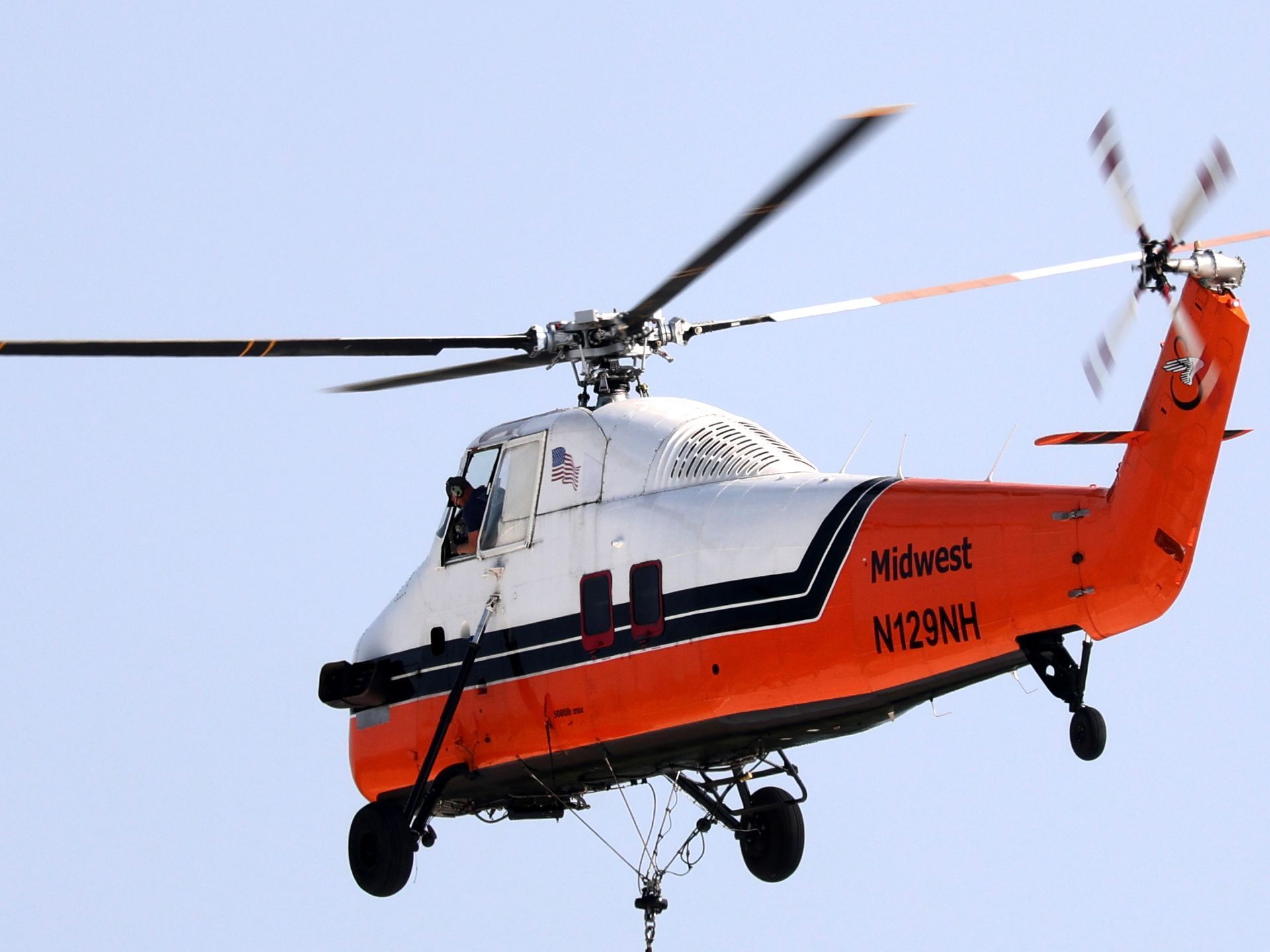 A white and orange Midwest helicopter flying against a clear sky, viewed from below with its landing gear visible.