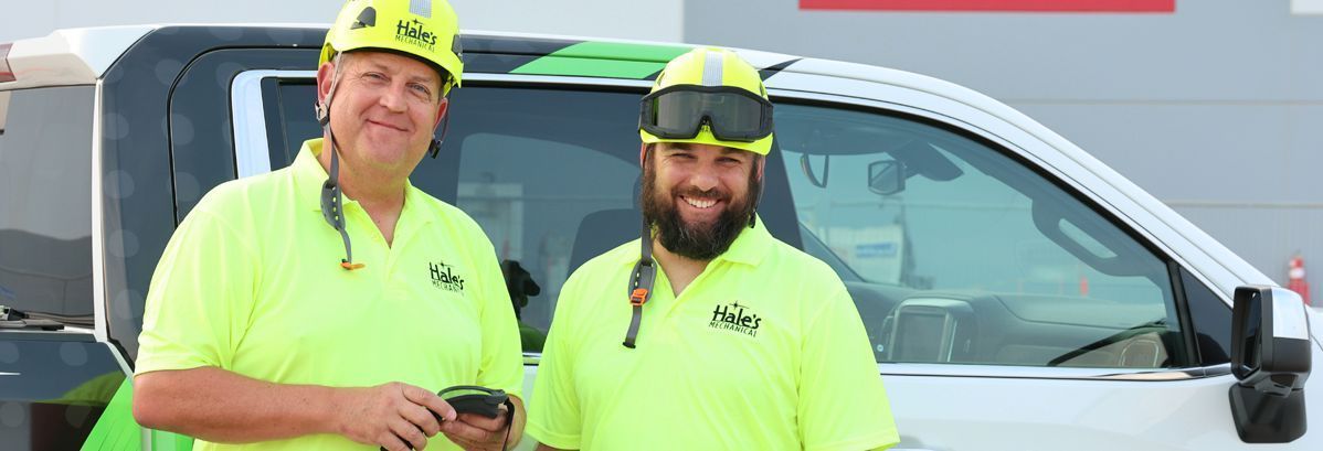 Two smiling workers in lime green high-visibility shirts and hard hats stand in front of a white service truck.